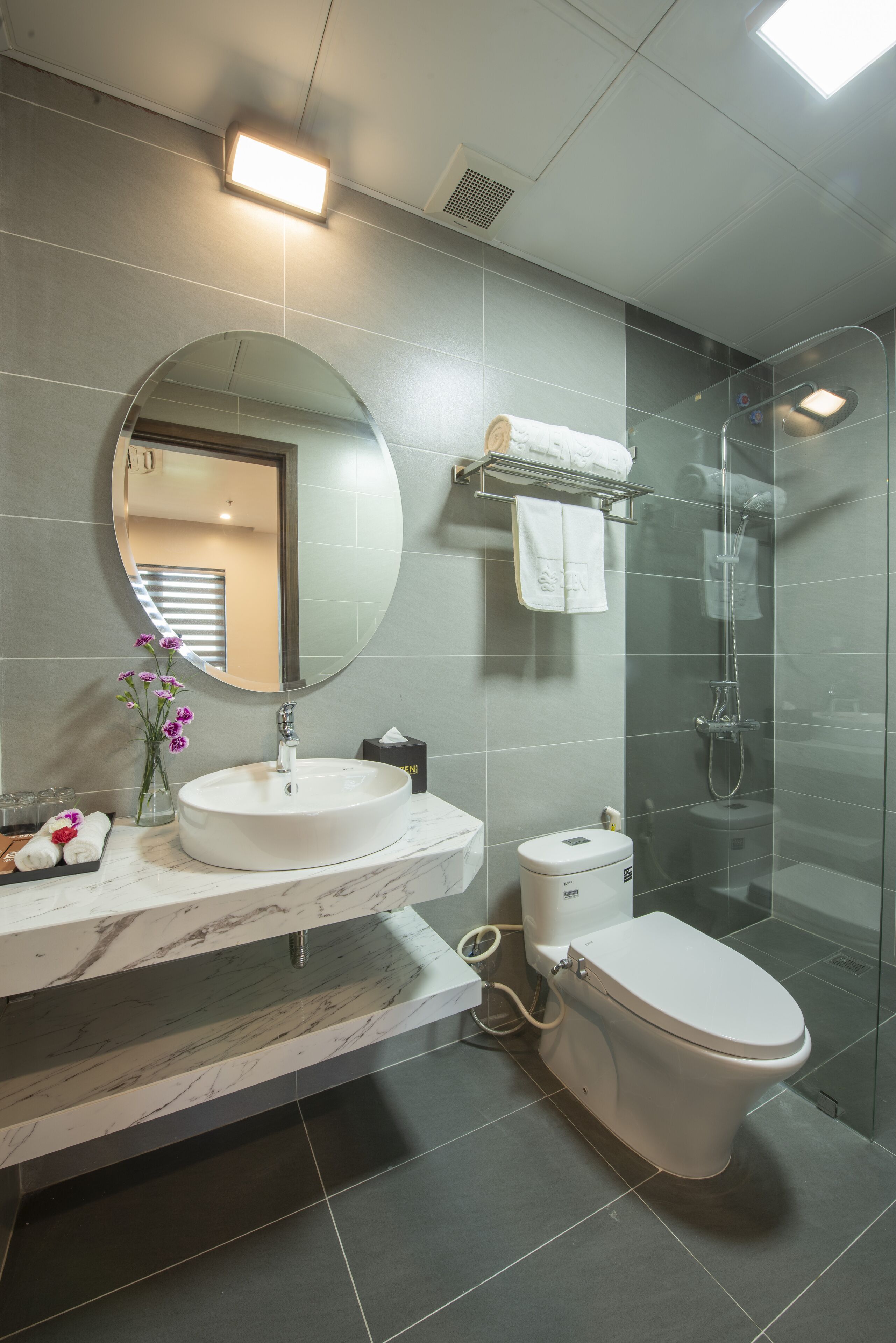 Een moderne badkamer met een ronde spiegel, een witte wastafel, een toilet en een douchecabine met glazen wanden.