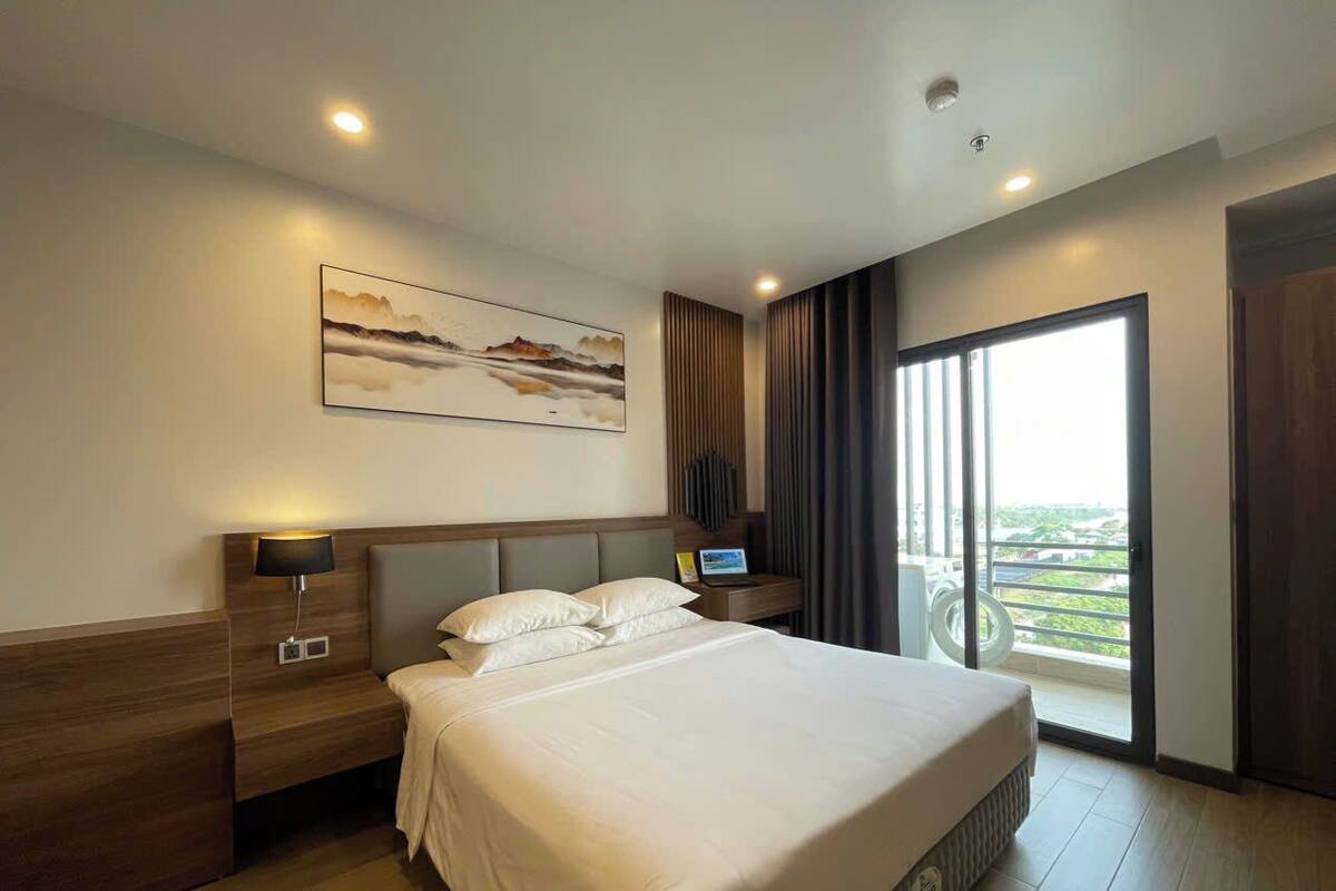 Ein Hotelzimmer mit einem großen Bett, einem Nachttisch mit Lampe, einem an der Wand montierten Fernseher und Blick ins Freie durch eine Schiebetür.