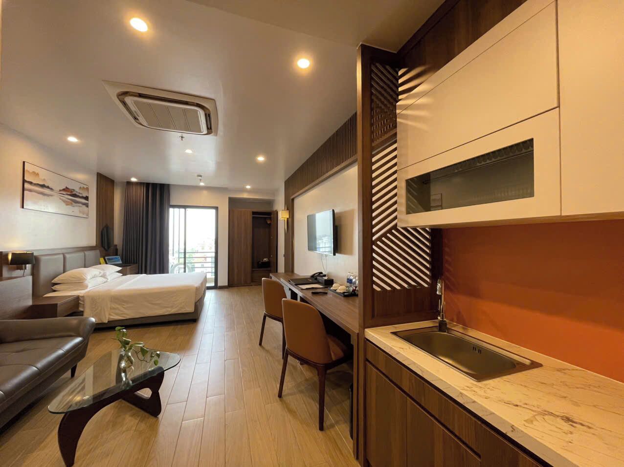 A modern hotel room with a bed, a sofa, a desk, a TV, and a kitchen area with a sink.