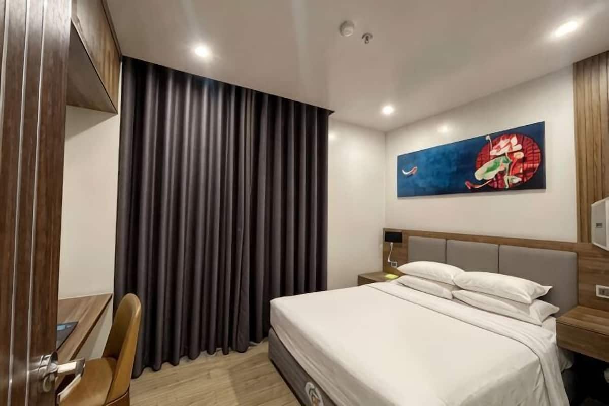 Ein Hotelzimmer mit Bett, Sessel, Schreibtisch, Nachttisch, einem Bild an der Wand und einem Vorhang.