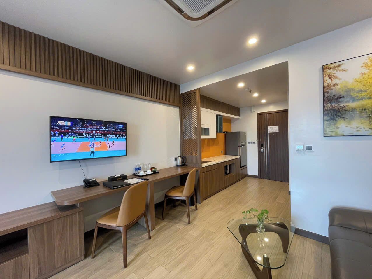 A modern hotel room with a TV, wooden furniture, a kitchen area, and a dining table.