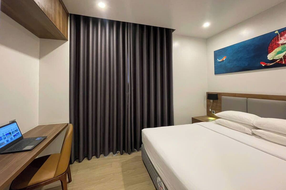 Ein Hotelzimmer mit einem Bett, einem Schreibtisch mit einem Laptop, einem Nachttisch und einem Gemälde an der Wand.