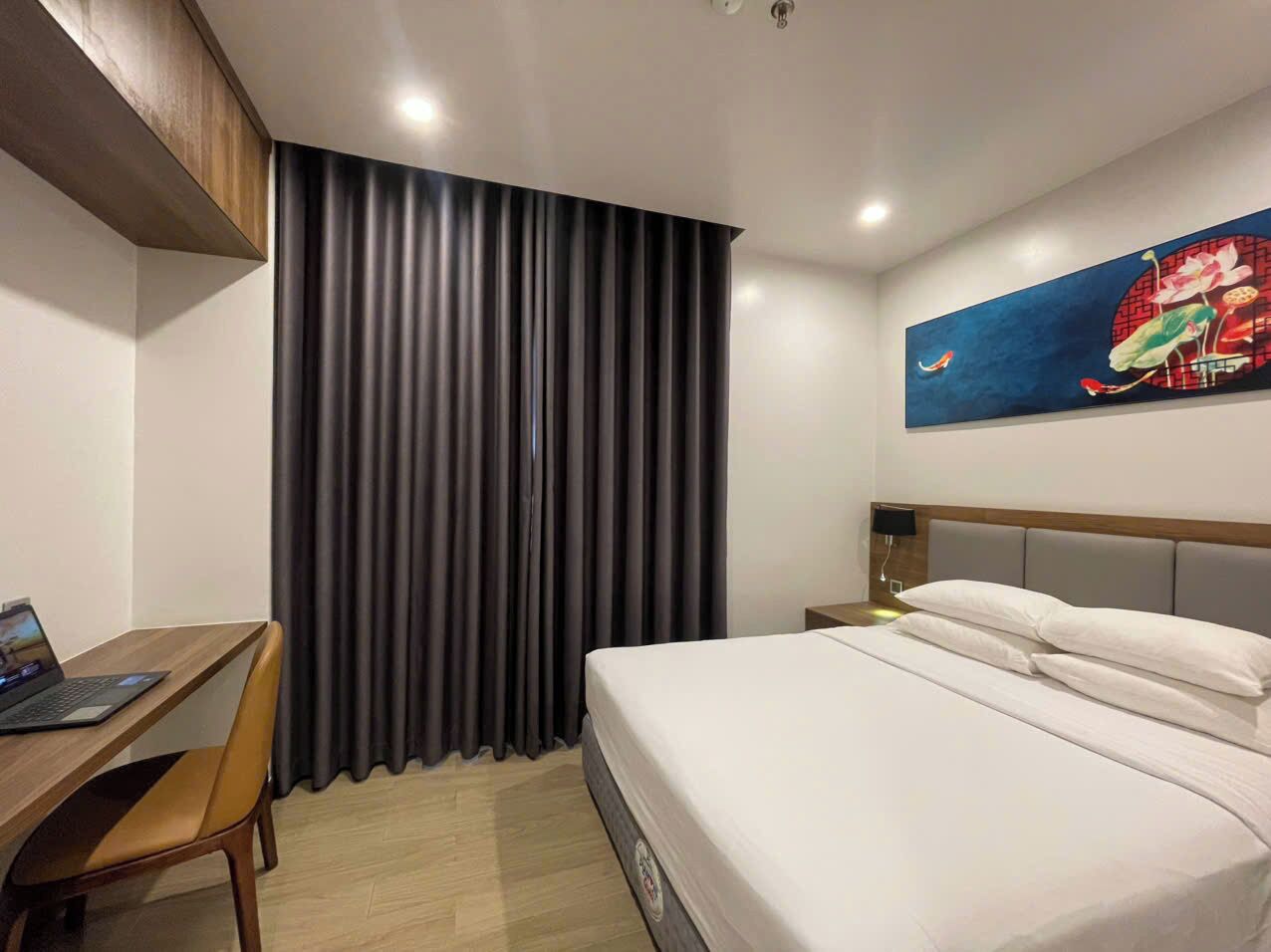 A hotel room with a bed, a desk with a laptop, a chair, a nightstand, and a painting on the wall.