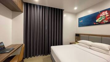 A hotel room with a bed, a desk with a laptop, a chair, a nightstand, and a painting on the wall.