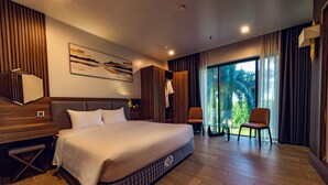 A modern hotel room with a large bed, wooden flooring, a view of greenery through the window, and a seating area with a chair and table. - Zen Riverside Hotel & Residences (Hai Phong)