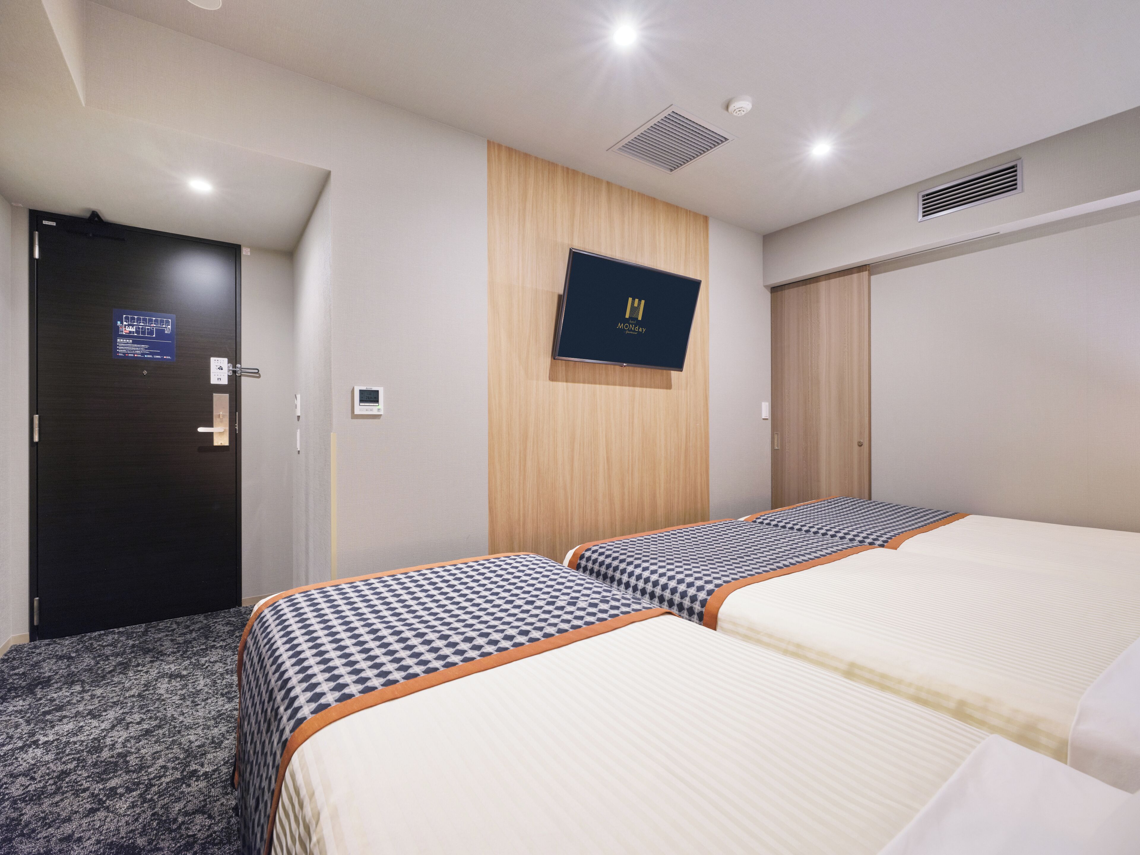 A hotel room with two beds, a wooden panel wall, a mounted TV, and a door with a map and information sign.