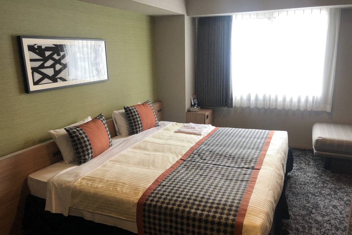 A hotel room with a large bed, a bedside table, a chair, a window with curtains, and a wall art piece.