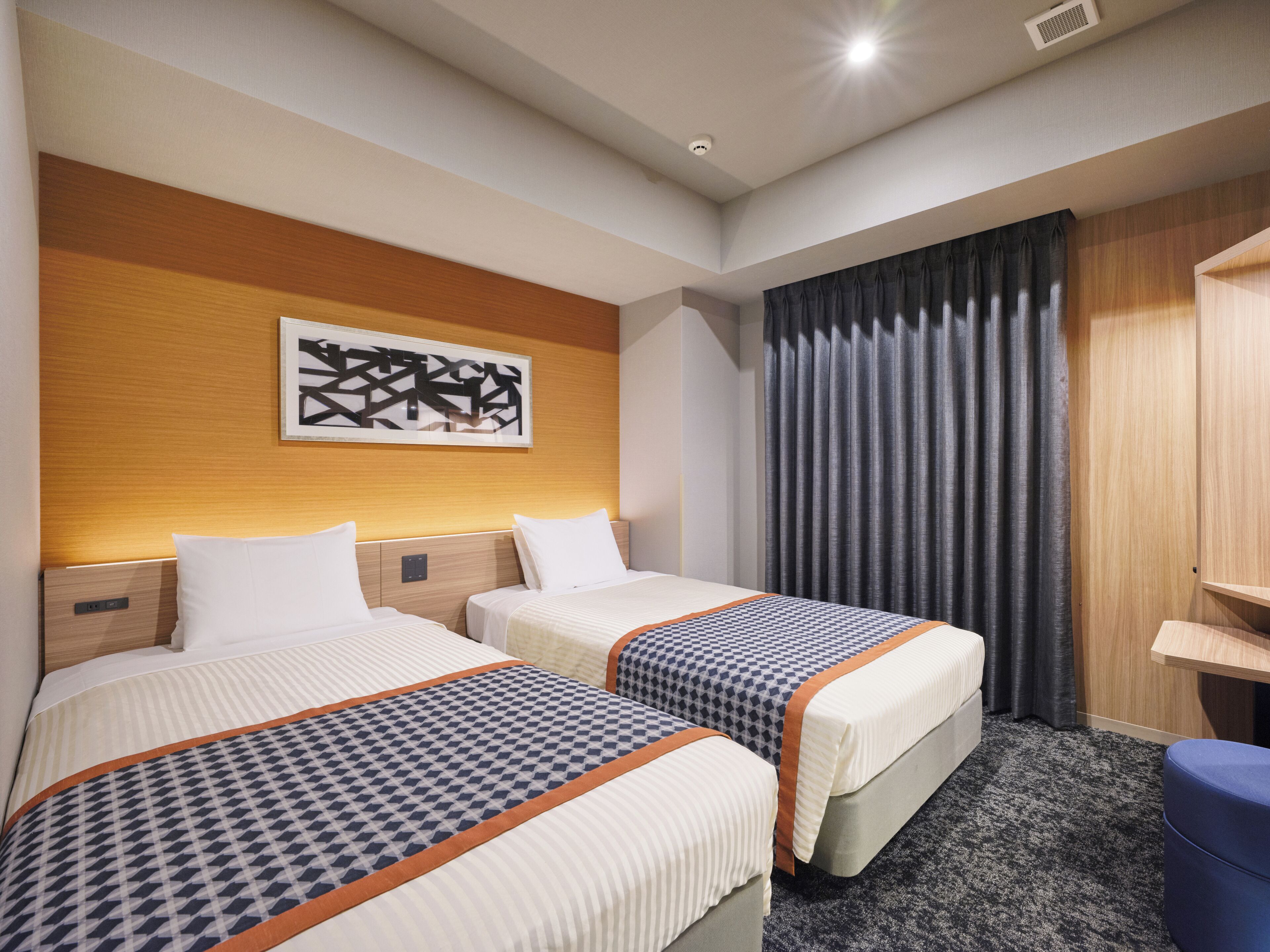 Habitación de hotel con dos camas, un escritorio, una silla, un ventanal con cortinas y una obra de arte colgada en la pared.