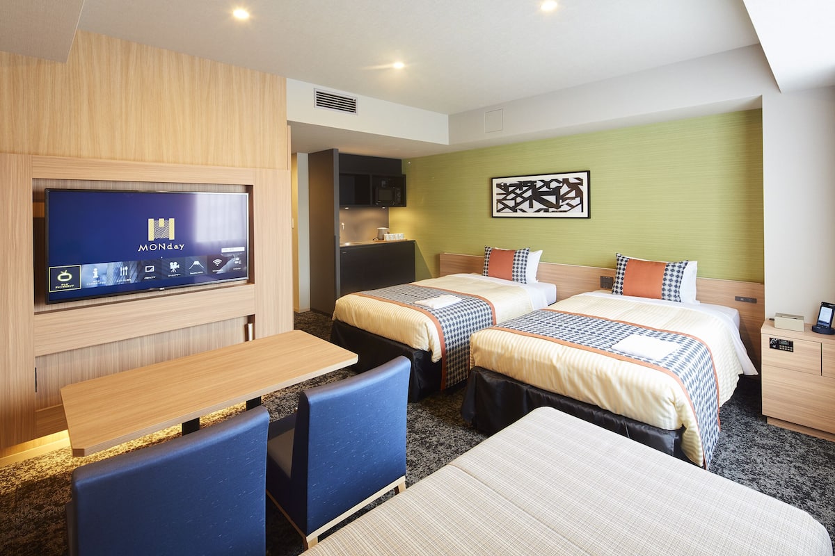 A hotel room with two beds, a TV, a desk, and a kitchenette.