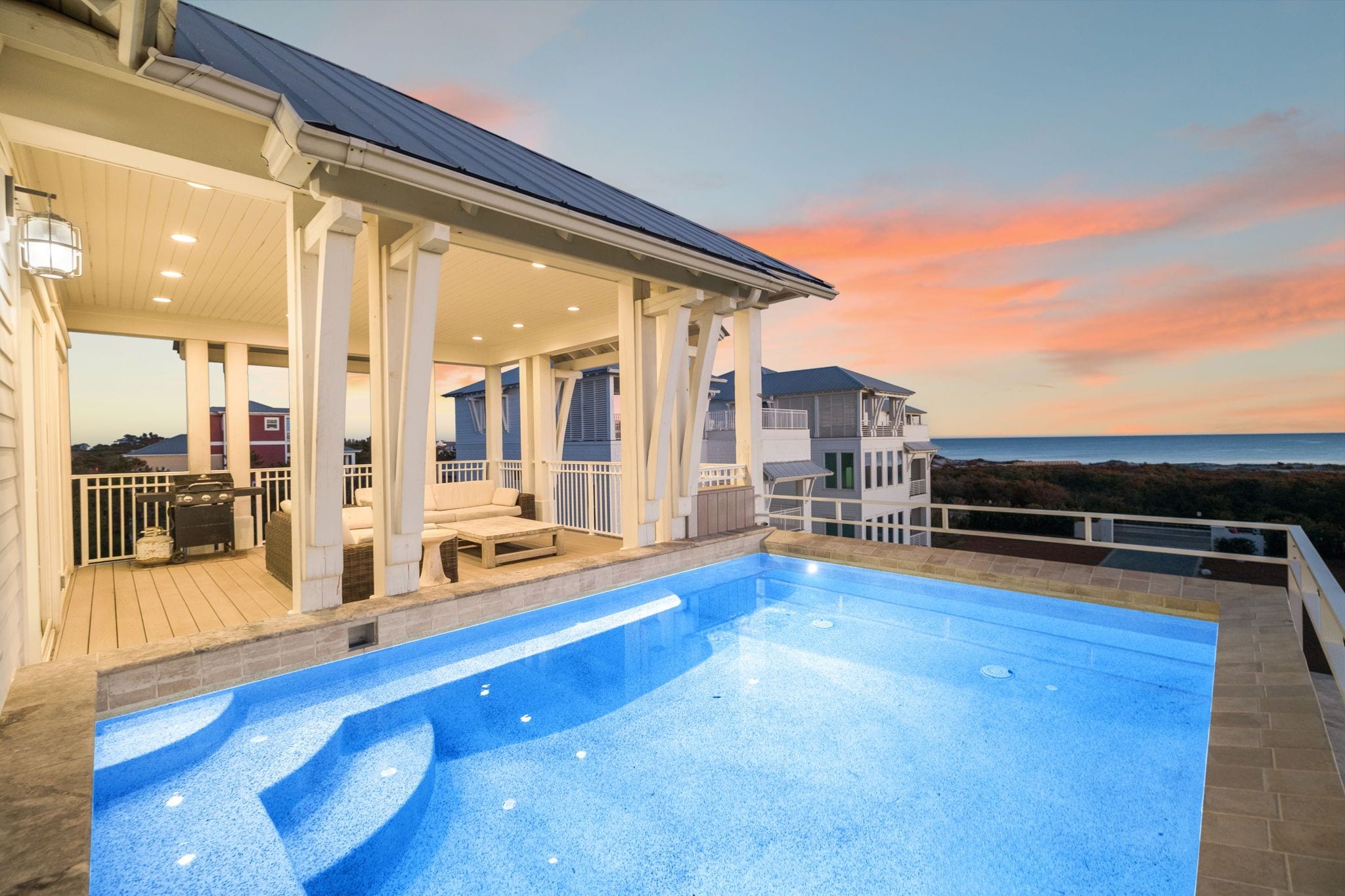 A swimming pool with a view of the ocean at sunset, a covered patio area, and a separate building with a balcony.