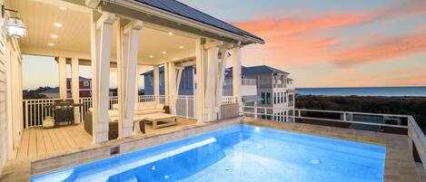 Una piscina con vistas al océano al atardecer, un área de patio techada y un edificio separado con balcón.
