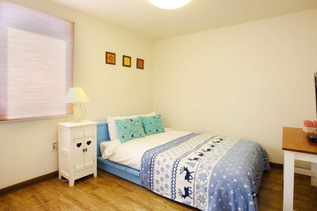 A bedroom with a bed, a nightstand, a lamp, a window with blinds, and a wall with three framed pictures.