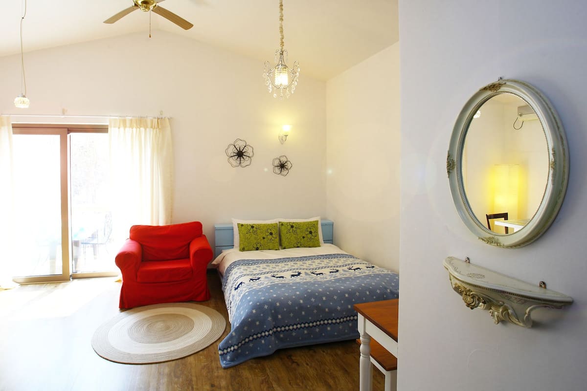 A bedroom with a bed, a red chair, a round rug, a chandelier, and a wall-mounted mirror.