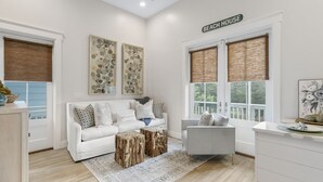 Un salon moderne avec un canapé blanc, un fauteuil gris et une table basse agrémentée de petites tables d’appoint en bois. Deux grandes fenêtres, équipées de stores bruns, et des œuvres d’art ornent les murs.