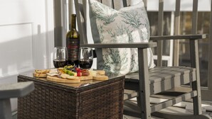 Une table en osier, avec une bouteille de vin, des verres et diverses collations, sur un balcon agrémenté d’une chaise et d’un coussin décoratif.