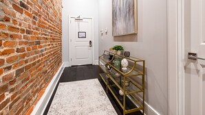 Un pasillo con pared de ladrillo, una puerta blanca, una consola dorada con objetos decorativos y un cuadro en la pared.