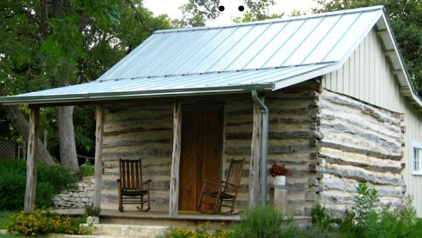 A rustic log cabin with a metal roof, a porch with two rocking chairs, and a stone wall foundation. - Butler Cabin at The Homestead B&B  (Dripping Springs,)