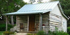 Une cabane rustique en rondins de bois, avec un toit en métal, une véranda dotée de deux fauteuils à bascule et des fondations en pierres.