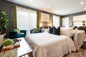 Een moderne hotelkamer met een groot bed, een zithoek met een blauwe bank en fauteuils, een eettafel met dienblad, en een groot raam met gordijnen.