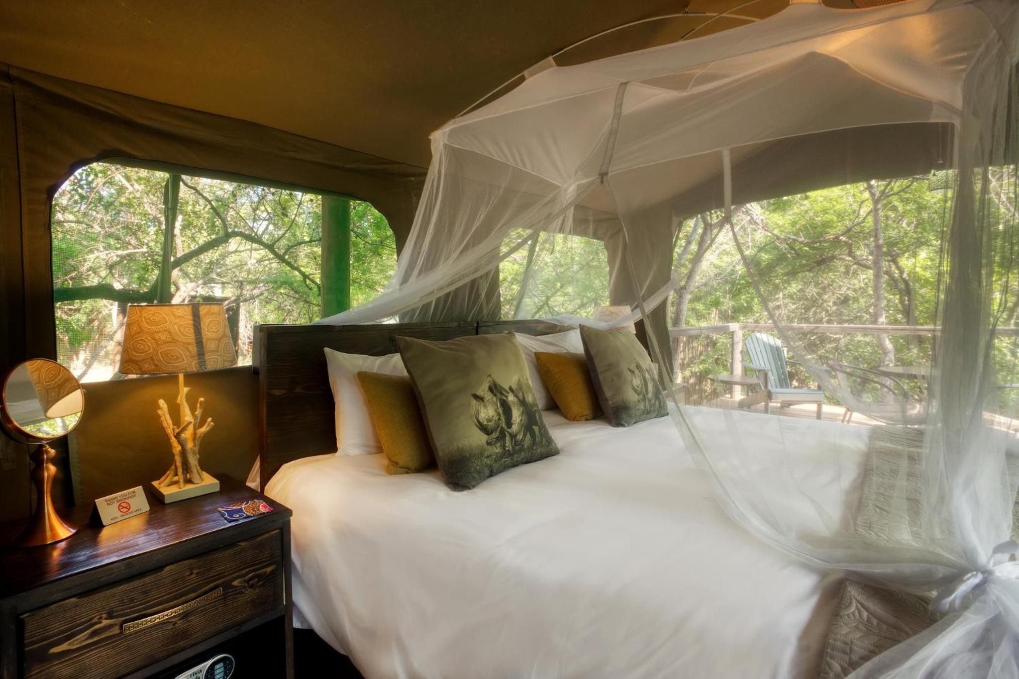 A bed with a canopy, a bedside table with a lamp, and a view of trees through a window.