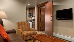A living room with a sofa, armchair, and coffee table. A TV is mounted on the wall, and a kitchen is visible through an open door. - Grand Lodge Peak 7 Suite (Breckenridge)