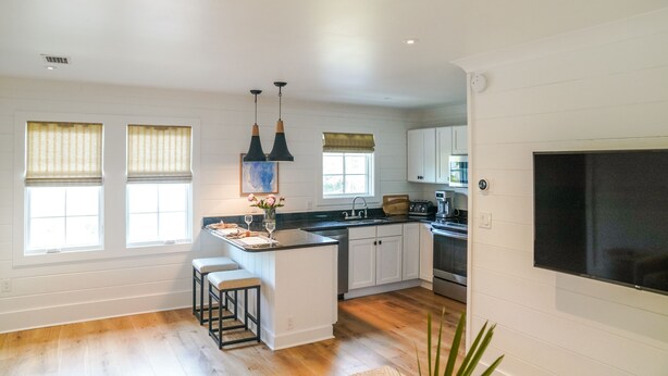 A modern kitchen with white cabinetry, a central island with stools, pendant lighting, and a large flat-screen TV mounted on the wall. - Home Away from Home! Beautiful Carriage House in Seagrove Close to the Beach (Santa Rosa Beach)