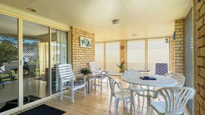 An outdoor patio with white chairs and a round table, a glass door leading outside, and a brick wall with a decorative pattern. - Sandy Paws - Burrum Heads- Walk to Beach -3BR- Aircon-Pool table (Burrum Heads)