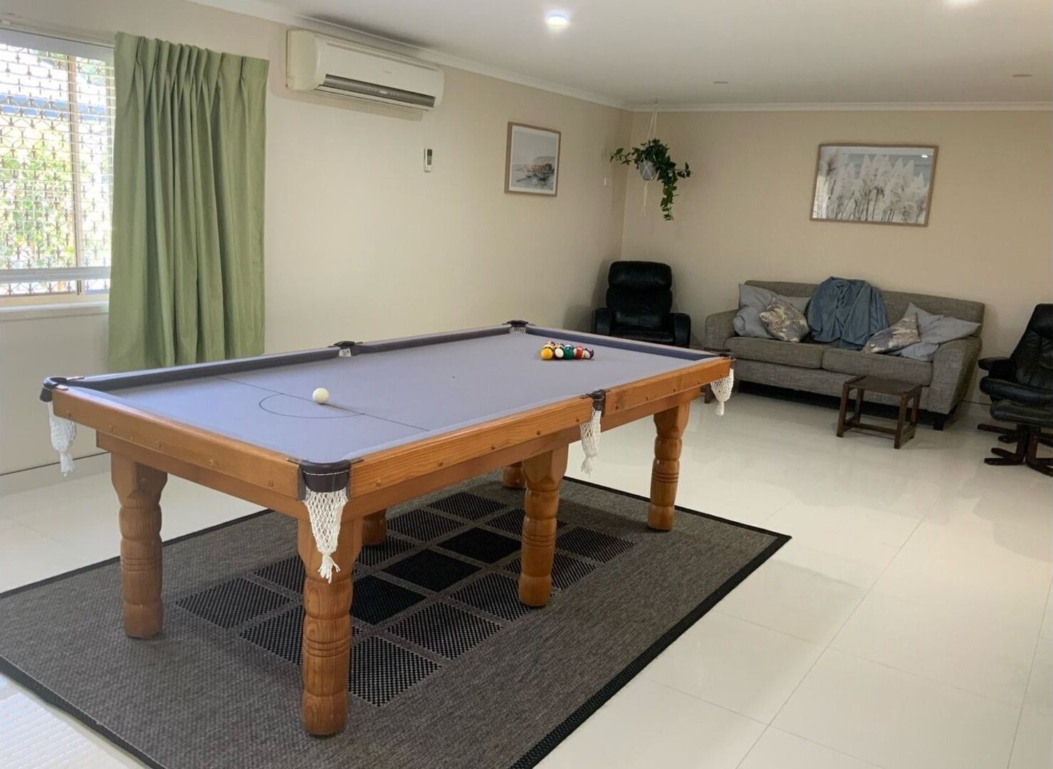 A pool table in a living room with a sofa and a chair, a window with curtains, and an air conditioner on the wall.