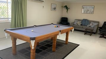 A pool table in a living room with a sofa and a chair, a window with curtains, and an air conditioner on the wall.