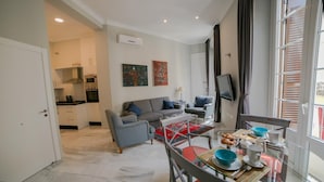 Una sala de estar moderna con un sofá, una mesa de comedor con sillas, una cocina al fondo y un ventanal con cortinas.