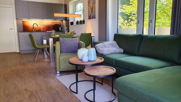 Ein modernes Wohnzimmer mit einem grünen Sofa, einem Essbereich mit Tisch und Stühlen sowie einer Küche im Hintergrund.