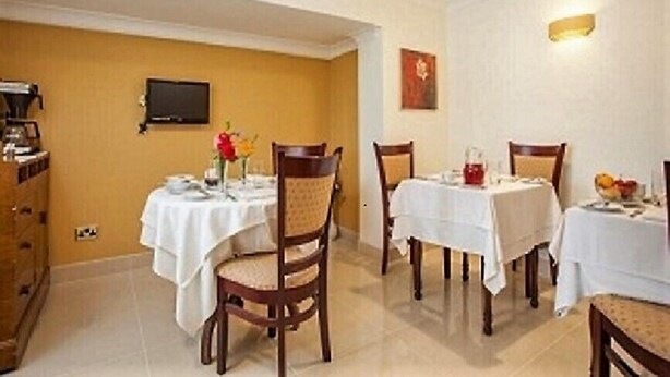 Un espace repas avec quatre tables, chacune recouverte d’une nappe blanche. Des chaises sont disposées autour des tables, et un téléviseur est fixé au mur.