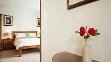 A hotel room with a bed, bedside table, and a vase with red flowers on a wooden nightstand.