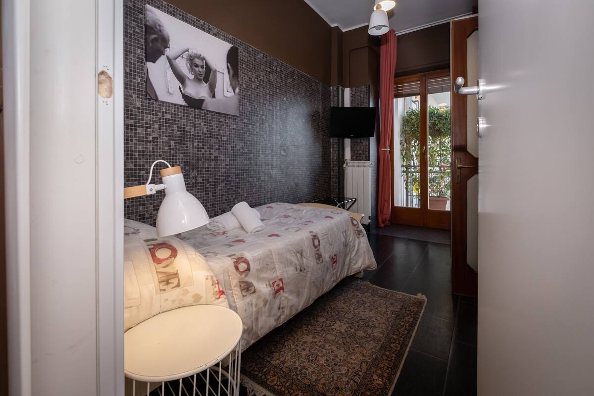 Une chambre à coucher comprenant un lit, une lampe de chevet, un téléviseur fixé au mur et une vue sur l’extérieur par une porte.