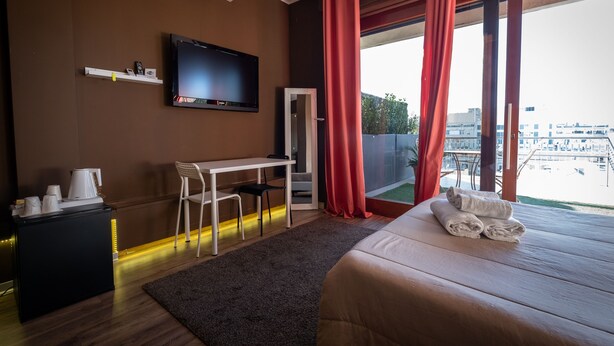 A hotel room with a bed, a desk, a chair, a TV, and a view of the city. - Dolci Emozioni (Bari)