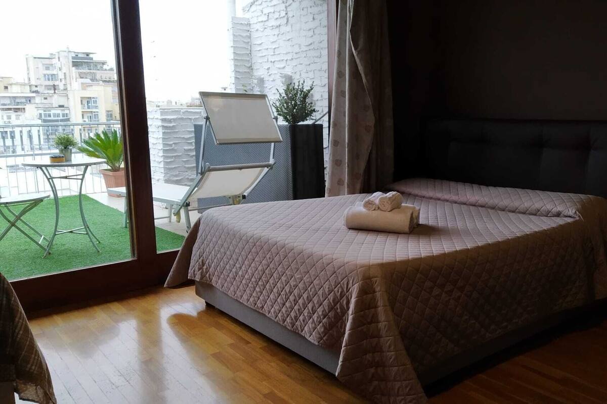 Une chambre avec un lit, une vue sur la ville, un balcon avec une table et des chaises, et une plante en pot.