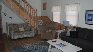 Ein Wohnzimmer mit Treppe, Sofa, Sessel, Couchtisch und zwei mit Vorhängen versehenen Fenstern.