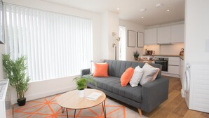 A modern living room with a grey sofa, orange cushions, a wooden coffee table, and a kitchen in the background. - Moonside - Stunning Angel Apartments (London)