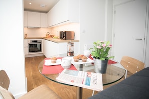 A modern kitchen with white cabinets, a microwave, and a coffee maker. There is a dining table with a glass top, a newspaper, and a potted plant with pink flowers.