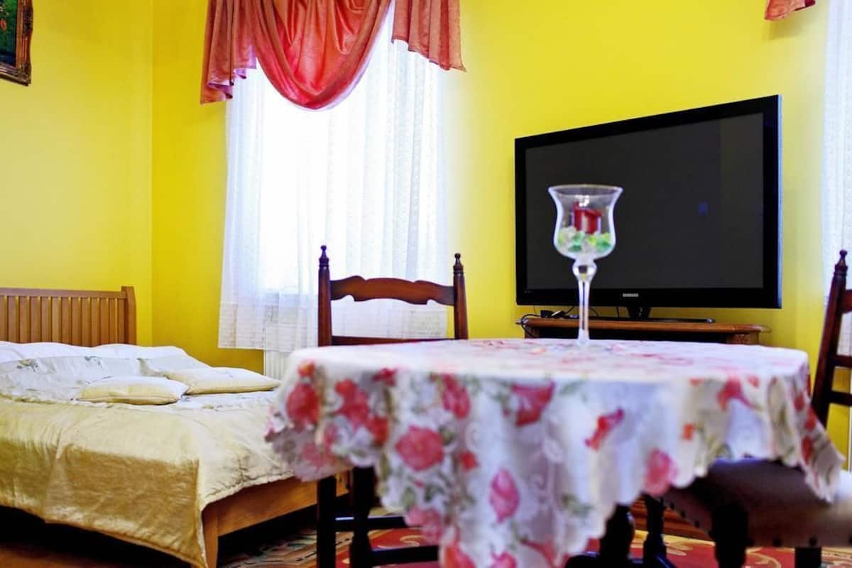A room with a bed, a table with a floral tablecloth, a television, and a window with curtains.