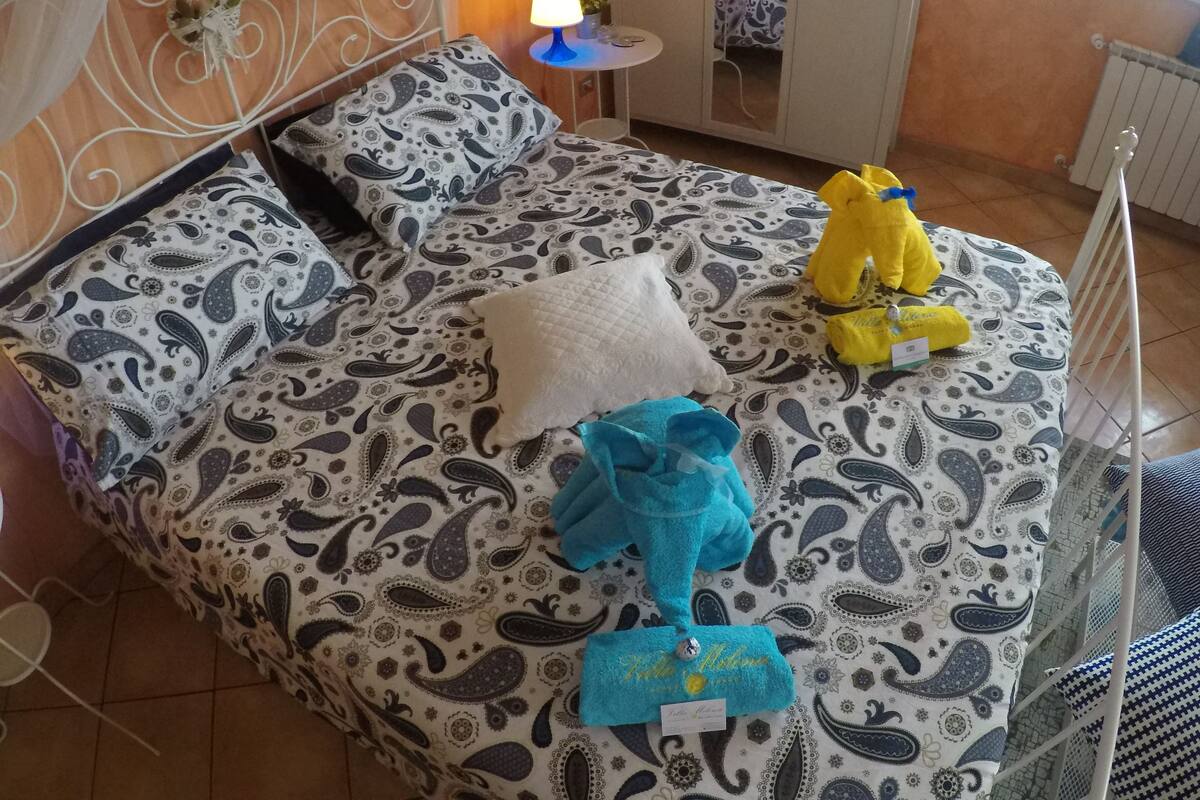 A bed with a patterned bedspread, pillows, and a stuffed toy. There is a small table with a lamp and a decorative item on the wall.
