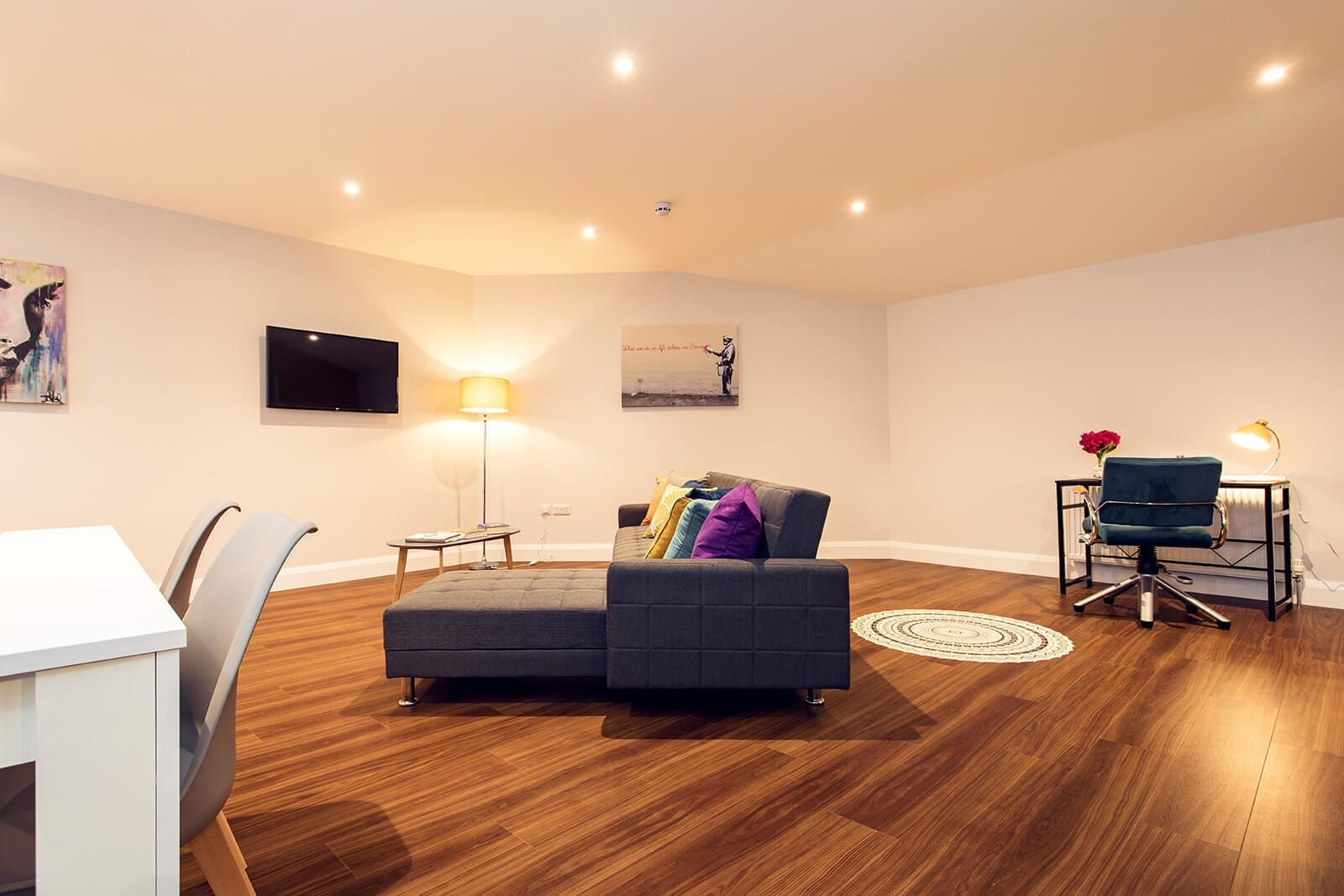 A modern living room with a grey sofa, a dining table with chairs, a TV mounted on the wall, and a floor lamp.