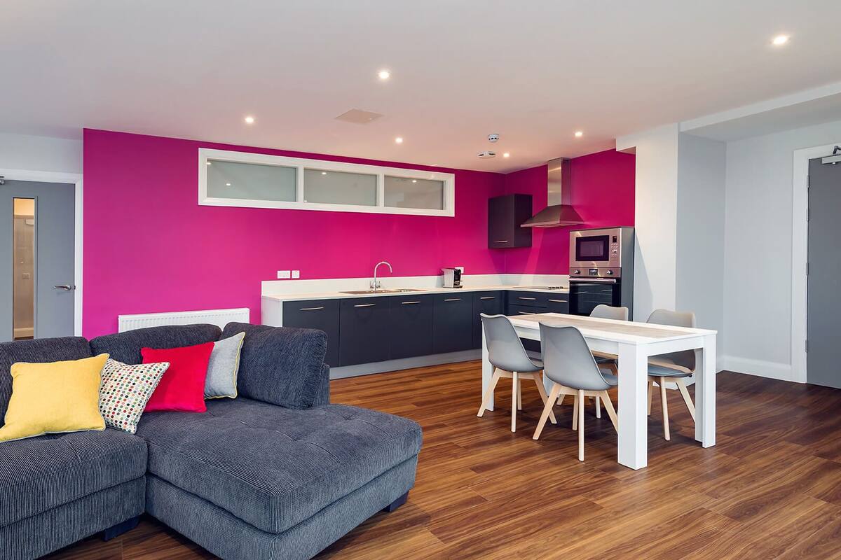 A modern apartment with a grey sofa, a dining table with chairs, a kitchen with pink walls, and a white ceiling.