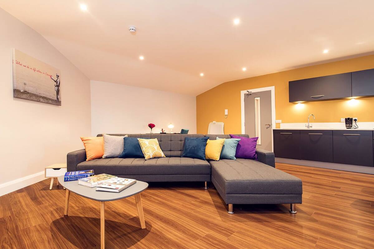 A modern living room with a grey sectional sofa, a coffee table, and a bookshelf. There is a kitchen area with dark cabinets and a white countertop.
