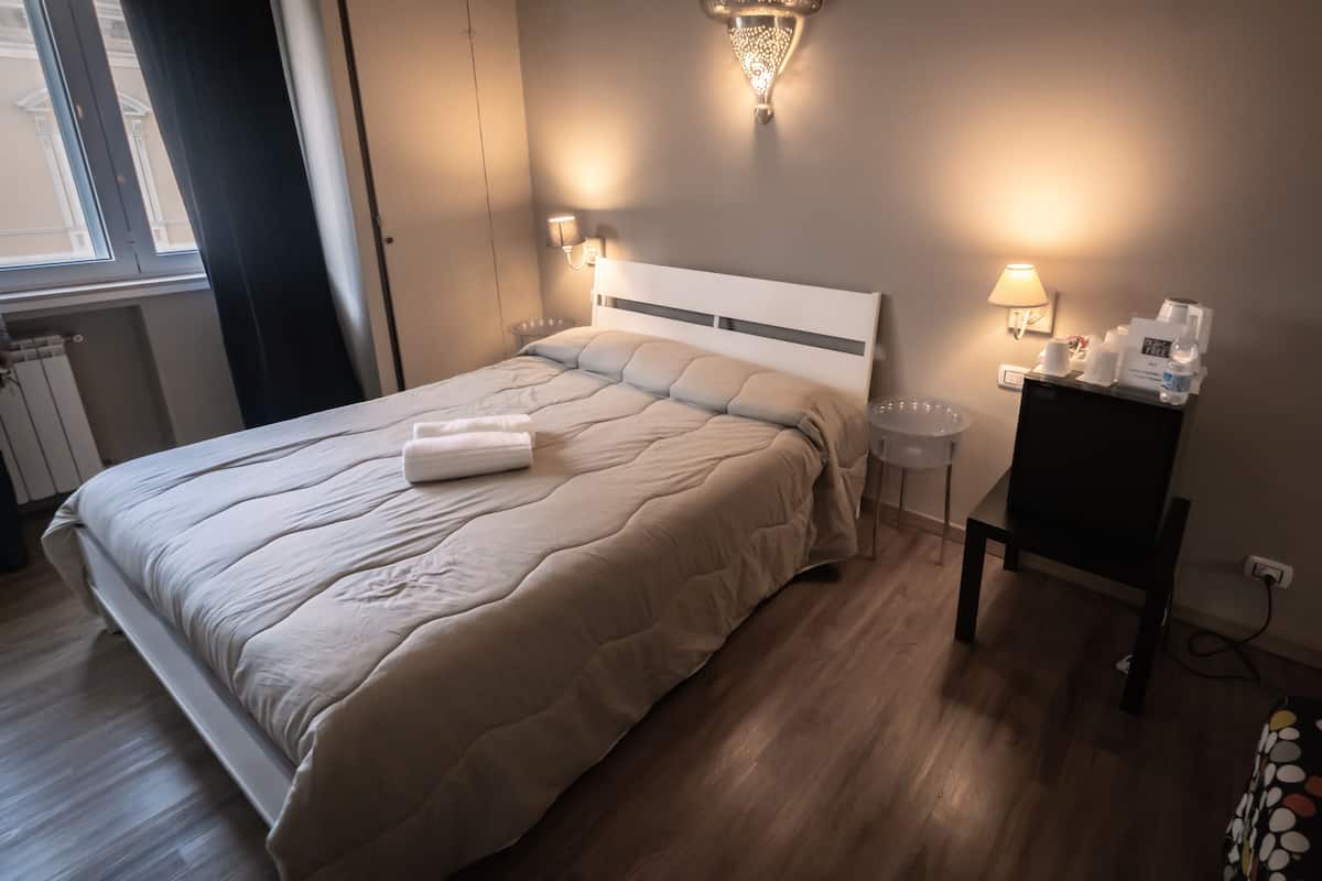 En snyggt bäddad säng med ett vitt överkast och en hopvikt handduk, ett sängbord med en lampa och ett litet sidobord med en telefon och en kopp.