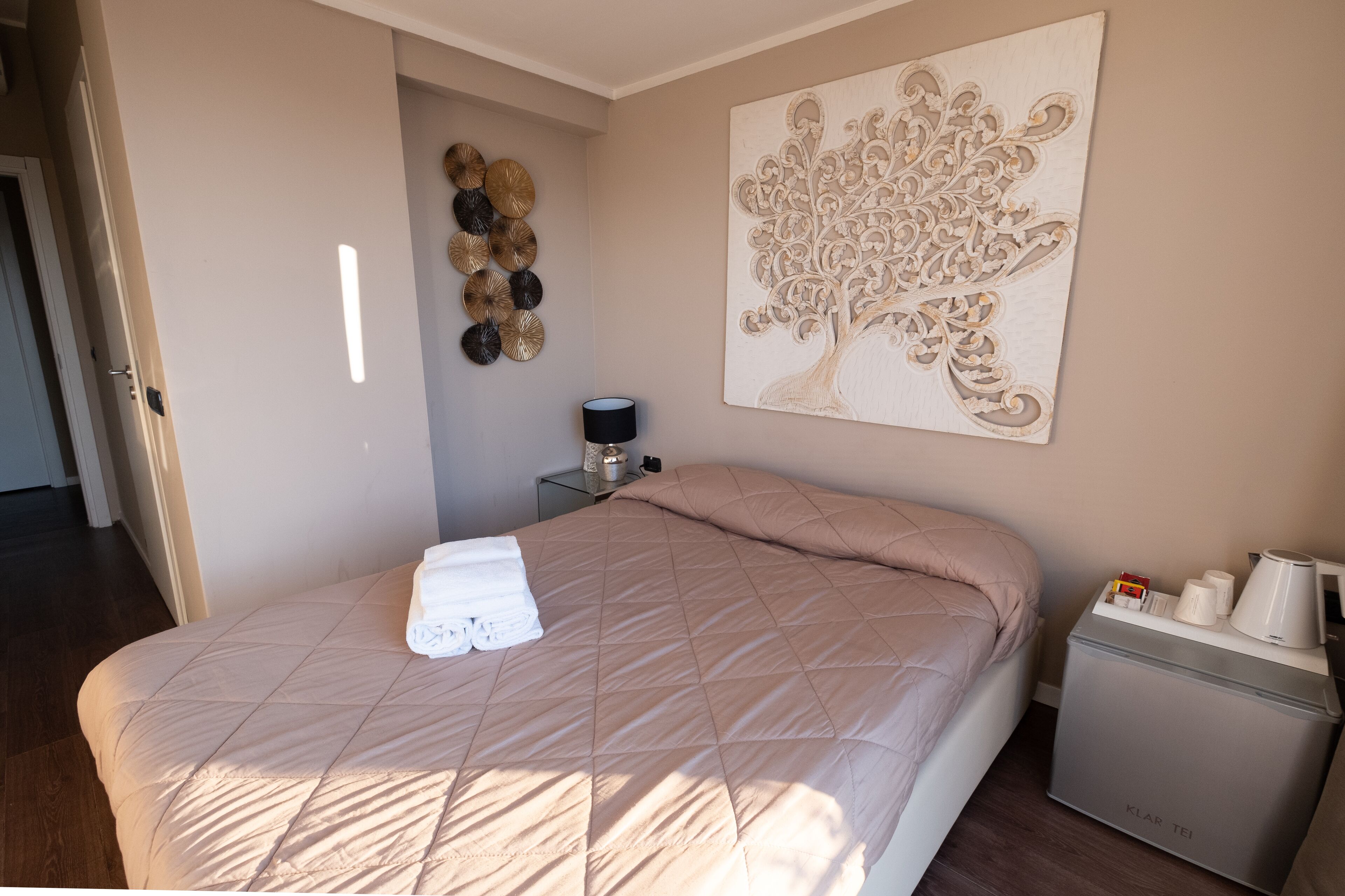 Una cama bien hecha con colcha color beige, una obra de arte de un árbol colgada en la pared, una mesita de noche con lámpara y un minibar.