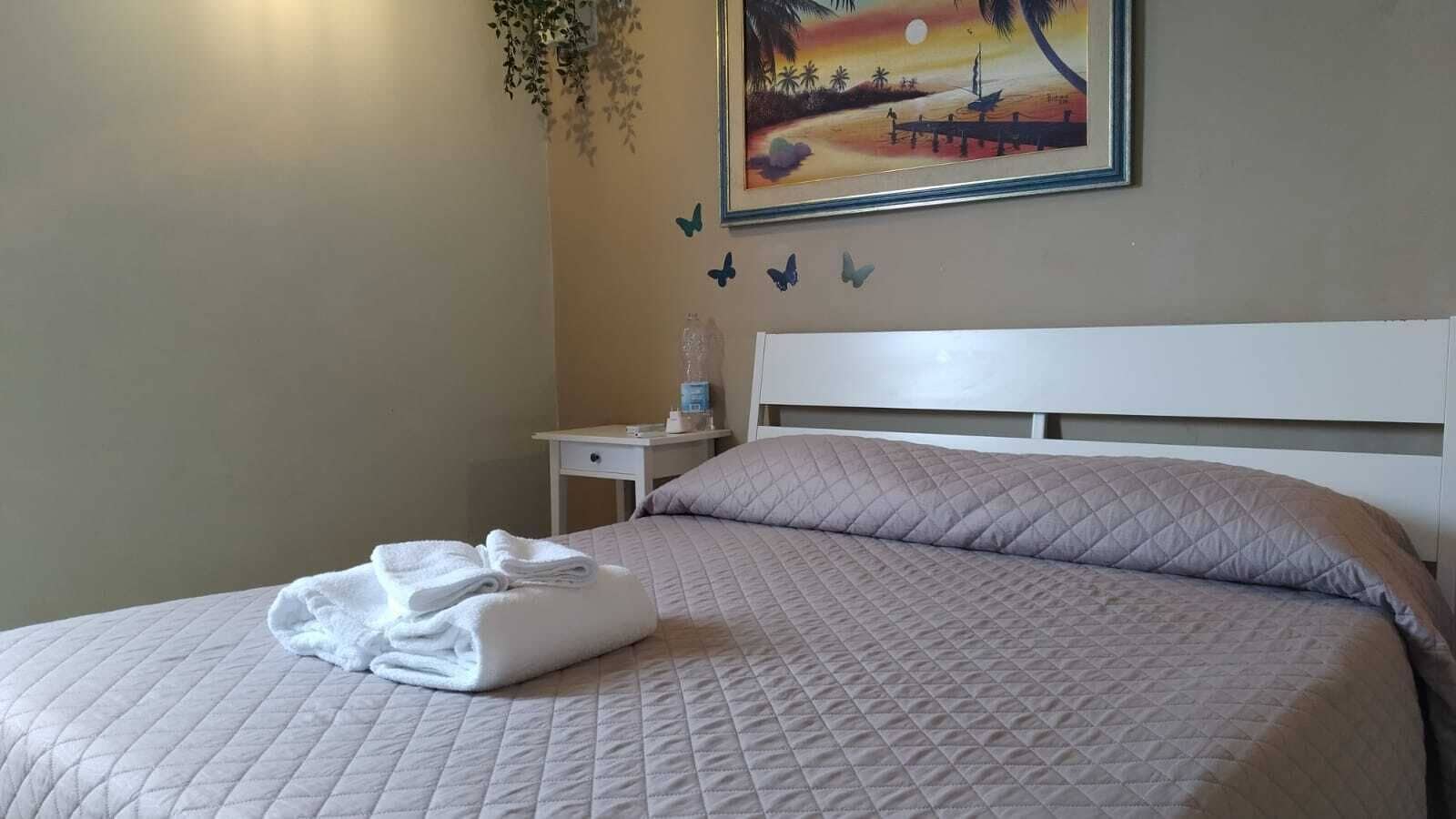 Una cama bien hecha con cubrecolcha acolchado y toallas dobladas, una mesita de noche con lámpara y una pared decorada con un cuadro de atardecer y motivos de mariposas.