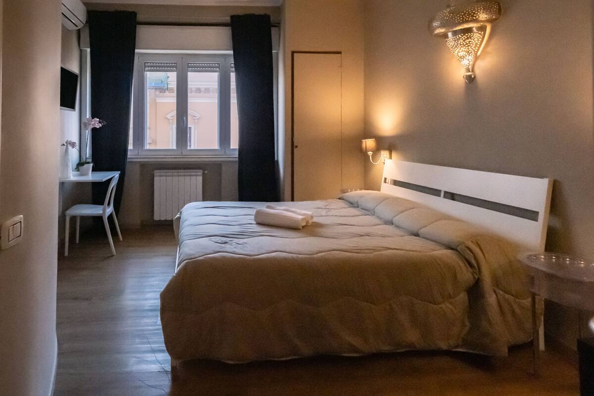 Ett hotellrum med en säng, ett skrivbord, en stol, ett fönster med gardiner och en väggmonterad lampa.