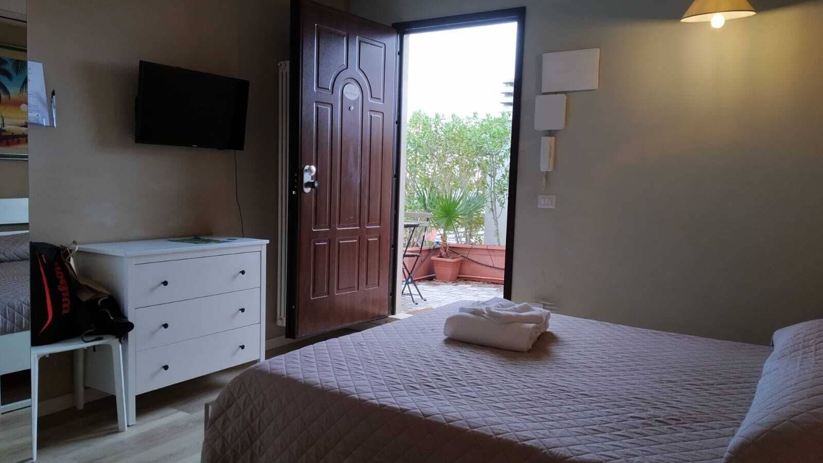 Een slaapkamer met een bed, een dressoir, een televisie en een deur naar een balkon met planten.