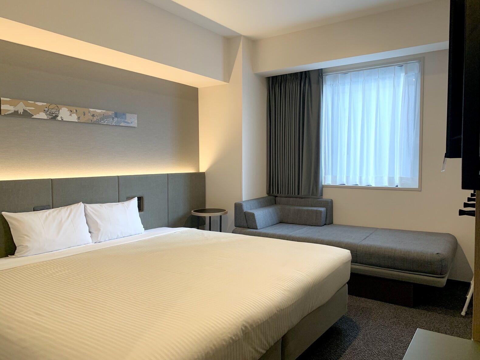 A hotel room with a large bed, a smaller sofa, a bedside table, a window with curtains, and a wall-mounted artwork.